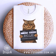 Футболка поганий кіт арешт, біла, зображена зверху, на фоні плетеного кошика, принт з котом у поліцейській табличці з написом 'BAD CAT'.