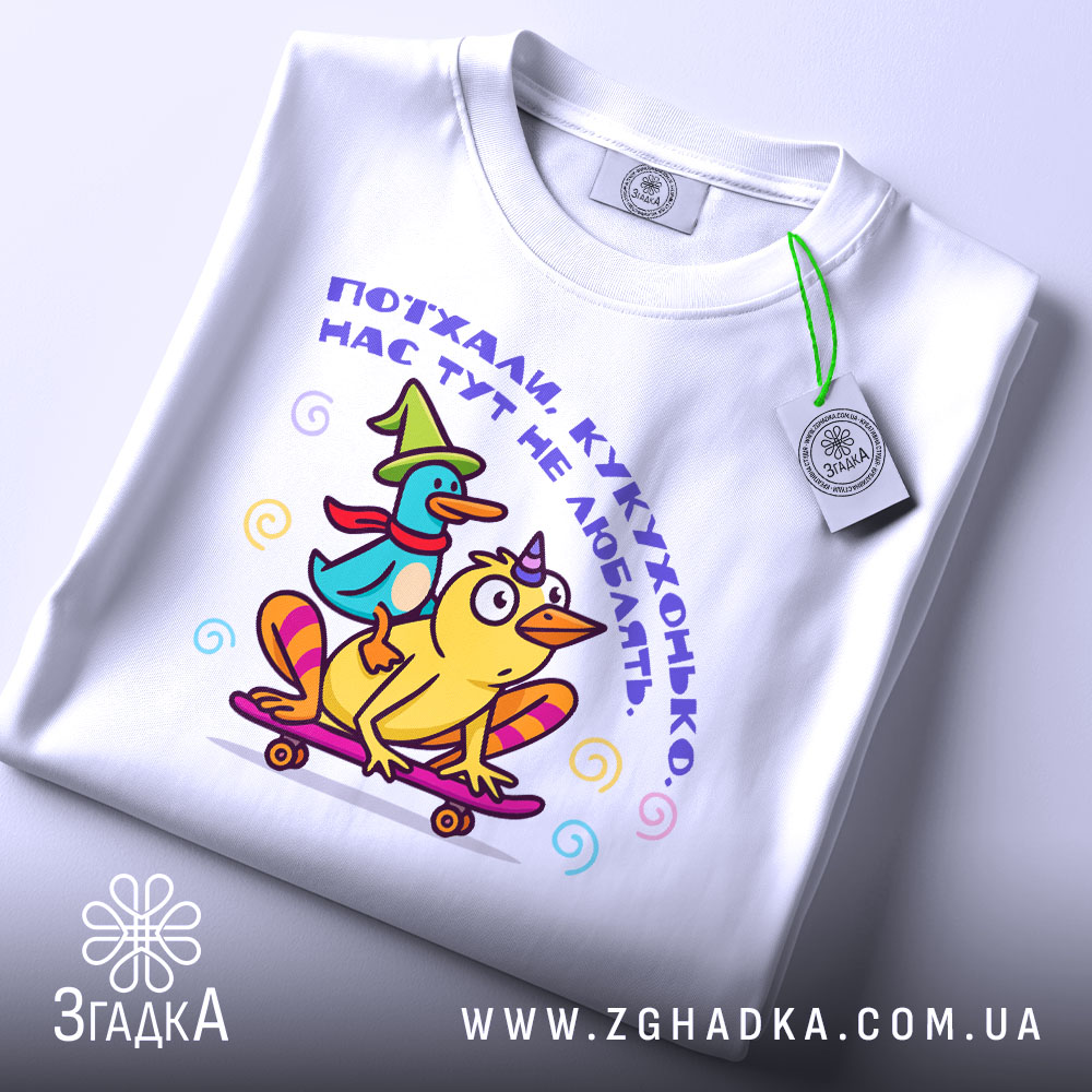 Футболка поїхала кукуха. Біла футболка, складена на столі, з яскравим принтом качок на скейтборді. Принт містить текст українською мовою. На футболці є етикетка з зеленим шнурком. Фон світло-сірий, що робить акцент на футболці.
