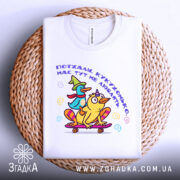 Футболка поїхала кукуха. Біла футболка, складена на плетеному кошику, з яскравим принтом качок на скейтборді. Принт містить текст українською мовою. Фон світло-сірий, що контрастує з текстурою кошика.