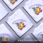 Футболка поїхала кукуха. Біла футболка на манекені, з принтом, де зображені дві качки на скейтборді. Принт яскравий, з жовтою та зеленою качкою, а також текстом українською мовою. Фон світло-сірий, що підкреслює кольори принта.