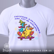 Футболка поїхала кукуха. Біла футболка, складена на столі, з яскравим принтом качок на скейтборді. Принт містить текст українською мовою. На футболці є етикетка з зеленим шнурком. Фон світло-сірий, що робить акцент на футболці.