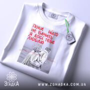 Футболка поки ніхто не бачить, біла, складена на поверхні, фон світлий, принт з двома фігурами в смугастих костюмах, текст червоним кольором: 'Поки ніхто не бачить Я досі тебе люблю', на футболці є етикетка з зеленим шнурком.