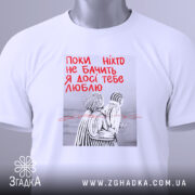 Футболка поки ніхто не бачить, біла, складена на поверхні, фон світлий, принт з двома фігурами в смугастих костюмах, текст червоним кольором: 'Поки ніхто не бачить Я досі тебе люблю', на футболці є етикетка з зеленим шнурком.