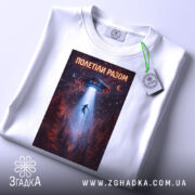 Футболка полетіли разом, білого кольору, складена на поверхні, фон світло-сірий, принт з космічним кораблем, що світить на людину, оточену лісом, з етикеткою на зеленому шнурку.