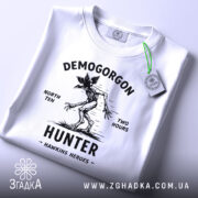 Футболка Полювання на Демогоргона білого кольору, складена на поверхні, з видимим етикеткою зеленого кольору, фон світло-сірий, принт чорного кольору з демогоргоном, текстом 'NORTH TEN TWO HOURS' та 'HUNTER HAWKINS HEROES'.