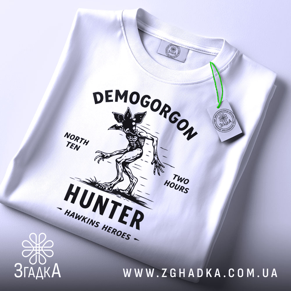 Футболка Полювання на Демогоргона білого кольору, складена на поверхні, з видимим етикеткою зеленого кольору, фон світло-сірий, принт чорного кольору з демогоргоном, текстом 'NORTH TEN TWO HOURS' та 'HUNTER HAWKINS HEROES'.