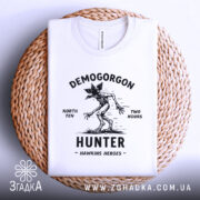 Футболка Полювання на Демогоргона білого кольору, складена на плетеній кошику, фон світло-сірий, принт чорного кольору з демогоргоном, текстом 'NORTH TEN TWO HOURS' та 'HUNTER HAWKINS HEROES'.