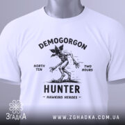 Футболка Полювання на Демогоргона білого кольору, складена на поверхні, з видимим етикеткою зеленого кольору, фон світло-сірий, принт чорного кольору з демогоргоном, текстом 'NORTH TEN TWO HOURS' та 'HUNTER HAWKINS HEROES'.