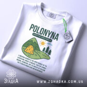 Футболка полонина карпати, біла, розкладена на поверхні, з видимою етикеткою, фон світлий, принт з малюнком гір, сонця та текстом про полонину.