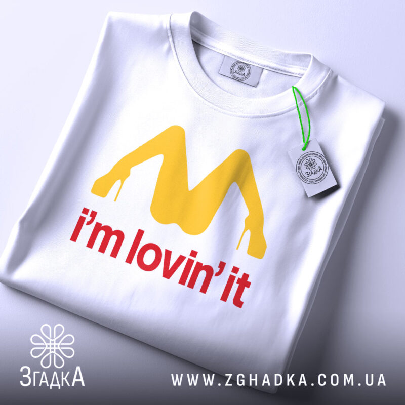 Футболка поп арт іронія, біла, складена на столі, фон світло-сірий, принт жовтого кольору у формі літери М з червоним написом 'i'm lovin' it', на футболці є етикетка з зеленим шнурком.