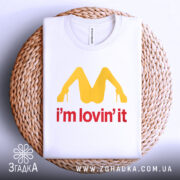 Футболка поп арт іронія, біла, складена на плетеній основі, фон світло-сірий, принт жовтого кольору у формі літери М з червоним написом 'i'm lovin' it'.
