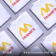 Футболка поп арт іронія, біла, зображена на манекені з правого боку, фон світло-сірий, принт жовтого кольору у формі літери М з червоним написом 'i'm lovin' it'.