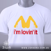 Футболка поп арт іронія, біла, складена на столі, фон світло-сірий, принт жовтого кольору у формі літери М з червоним написом 'i'm lovin' it', на футболці є етикетка з зеленим шнурком.