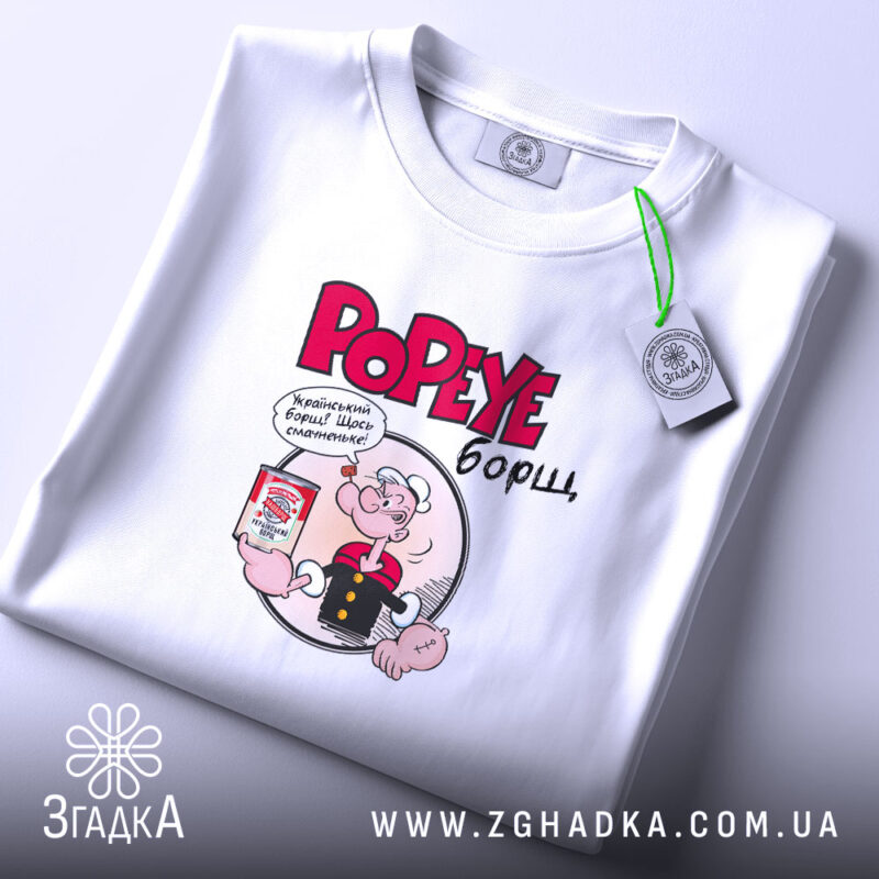 Футболка Popeye борщ, біла, складена на поверхні, фон світло-сірий, принт з Попаєм, який тримає банку борщу, текст червоним кольором, на комірці є етикетка.