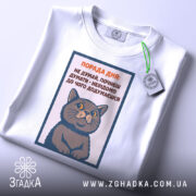 Футболка порада дня: Біла футболка з принтом кота, зображена зверху, фон світлий, на футболці видно текст поради дня, а також тег з логотипом бренду.