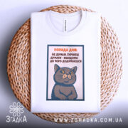 Футболка порада дня: Біла футболка з принтом кота, зображена зверху на плетеній основі, фон світлий, принт містить текст поради дня та малюнок кота, що сидить.
