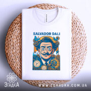 Футболка портрет Сальвадора Далі, біла, складена на плетеній кошику, фон світло-сірий, принт з портретом Далі, оточений метеликами та годинниками, з видимим ярликом на комірці.