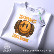 Футболка посміхайся, біла, складена на поверхні, з видимим етикеткою, фон світло-сірий, принт з усміхненим смайлом та черепами, текст "ПОСМІХАЙСЯ ПОКИ ЖИВИЙ" у жовтому кольорі.