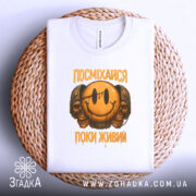 Футболка посміхайся, біла, складена на плетеній кошику, фон світло-сірий, принт з усміхненим смайлом та черепами, текст "ПОСМІХАЙСЯ ПОКИ ЖИВИЙ" у жовтому кольорі.