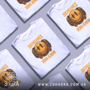 Футболка посміхайся, біла, зображена з фронтального ракурсу на манекені, фон світло-сірий, принт з усміхненим смайлом та черепами, текст "ПОСМІХАЙСЯ ПОКИ ЖИВИЙ" у жовтому кольорі.