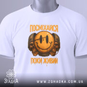 Футболка посміхайся, біла, складена на поверхні, з видимим етикеткою, фон світло-сірий, принт з усміхненим смайлом та черепами, текст "ПОСМІХАЙСЯ ПОКИ ЖИВИЙ" у жовтому кольорі.
