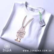 Футболка поверніть доброту, біла, складена на столі, фон світло-сірий, принт з малюнком кролика, що тримає табличку з написом 'Поверніть доброту в моду!', на комірці є етикетка.