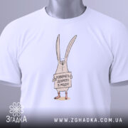 Футболка поверніть доброту, біла, складена на столі, фон світло-сірий, принт з малюнком кролика, що тримає табличку з написом 'Поверніть доброту в моду!', на комірці є етикетка.