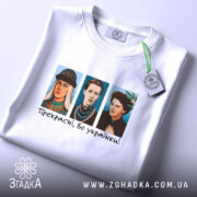 Футболка прекрасні бо українки. Біла футболка, зображена з верхнього ракурсу, на ній три персонажі в кольорових костюмах, текст "Прекрасні, бо українки!" під ними, на футболці є етикетка з зеленим шнурком, фон світлий.