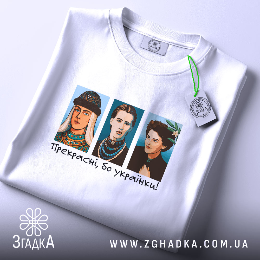 Футболка прекрасні бо українки. Біла футболка, зображена з верхнього ракурсу, на ній три персонажі в кольорових костюмах, текст "Прекрасні, бо українки!" під ними, на футболці є етикетка з зеленим шнурком, фон світлий.
