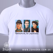 Футболка прекрасні бо українки. Біла футболка, зображена з верхнього ракурсу, на ній три персонажі в кольорових костюмах, текст "Прекрасні, бо українки!" під ними, на футболці є етикетка з зеленим шнурком, фон світлий.