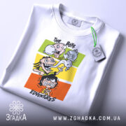 Футболка пригоди хлопців, біла, складена на столі, фон світлий, принт з чотирма персонажами на кольорових прямокутниках, з етикеткою на рукаві.