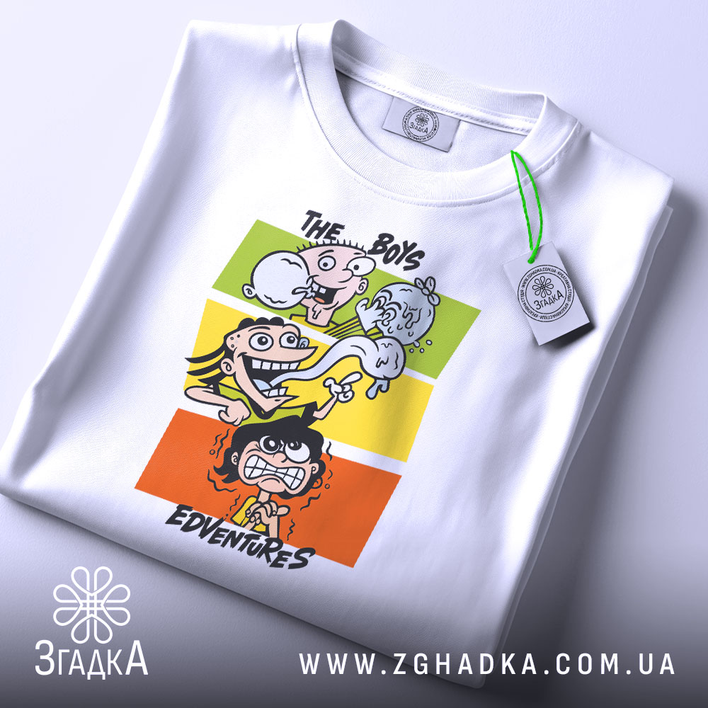 Футболка пригоди хлопців, біла, складена на столі, фон світлий, принт з чотирма персонажами на кольорових прямокутниках, з етикеткою на рукаві.