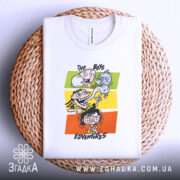 Футболка пригоди хлопців, біла, складена на плетеній кошику, фон світлий, принт з чотирма персонажами на кольорових прямокутниках, видно етикетку на горловині.
