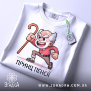 Футболка Принц Пенсії білого кольору, зображена зверху, на фоні світло-сірого кольору, принт з дідусем у червоній сорочці, палиця в руці, на футболці є етикетка.