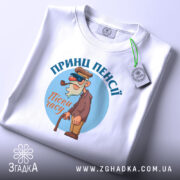 Футболка принц пенсії, білого кольору, складена на столі, фон світло-сірий, принт з мультяшним персонажем у капелюсі та окулярах, тримає тростину, текст 'Пісні часу' під персонажем, на футболці прикріплена етикетка.
