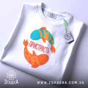 Футболка пристрасть риби. Складена біла футболка з принтом двох рибок, одна блакитна, інша оранжева. На футболці напис 'ПРИСТРАСТЬ'. Вид зверху, фон світло-сірий, з етикеткою на футболці.