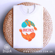 Футболка пристрасть риби. Складена біла футболка з принтом двох рибок на плетеній кошику. Рибки блакитного та оранжевого кольорів, напис 'ПРИСТРАСТЬ' розташований між ними. Фон білий, що підкреслює текстуру кошика.