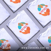 Футболка пристрасть риби. Біла футболка на манекені, з принтом двох рибок: одна блакитна з жовтими лусочками, інша оранжева з червоними відтінками. На футболці напис 'ПРИСТРАСТЬ'. Фон світло-сірий, що підкреслює яскравість кольорів принта.