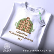 Футболка психіатрична лікарня, біла, складена на столі, фон світло-сірий, принт з будівлею лікарні в зеленому колі, текст під принтом: 'ОБЛАСНА ПСИХІАТРИЧНА ЛІКАРНЯ №2', на комірці видно етикетку.