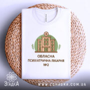 Футболка психіатрична лікарня, біла, складена на плетеному кошику, фон світлий, принт з будівлею лікарні в зеленому колі, текст під принтом: 'ОБЛАСНА ПСИХІАТРИЧНА ЛІКАРНЯ №2'.