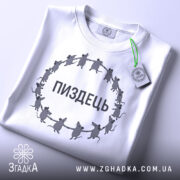 Футболка пиздець, біла, складена на поверхні, фон світло-сірий, принт з мишами, що тримаються за руки, текст "ПИЗДЕЦЬ" великими літерами, з етикеткою зеленого кольору.