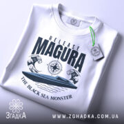 Футболка Release Magura білого кольору, зображена зверху, на ній прикріплена етикетка зеленого кольору, фон світлий, принт з човном та хвилями, текст 'THE BLACK SEA MONSTER'.