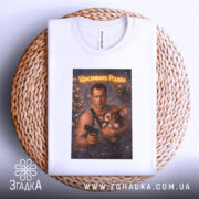 Футболка різдвяний бойовик, біла, складена на плетеній кошику, фон нейтральний, принт зображує чоловіка з пістолетом та істотою, текст на принті 'Щасливого Різдва'.