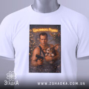 Футболка різдвяний бойовик, біла, складена на поверхні, фон світлий, принт зображує чоловіка з пістолетом та істотою, текст на принті 'Щасливого Різдва', на футболці є етикетка з зеленим шнурком.