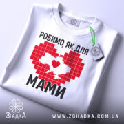 Футболка робимо як для мами, біла, зображена зверху на плоскій поверхні, фон світло-сірий, принт червоного кольору у формі серця з контуром України, напис чорним шрифтом, на комірці є етикетка.