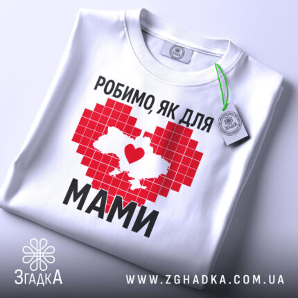 Футболка робимо як для мами, біла, зображена зверху на плоскій поверхні, фон світло-сірий, принт червоного кольору у формі серця з контуром України, напис чорним шрифтом, на комірці є етикетка.