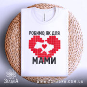 Футболка робимо як для мами, біла, зображена на плоскій поверхні, фон світло-сірий, принт червоного кольору у формі серця з контуром України, напис чорним шрифтом, футболка розташована на плетеній основі.