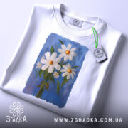Футболка ромашки, білого кольору, зображена з верхнього ракурсу, складена на столі, фон світло-сірий, принт з трьох ромашок на блакитному фоні, з етикеткою на комірці.