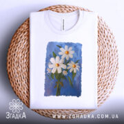 Футболка ромашки, білого кольору, складена на плетеній кошику, фон світло-сірий, принт з трьох ромашок на блакитному фоні, з зеленим листям.