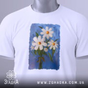 Футболка ромашки, білого кольору, зображена з верхнього ракурсу, складена на столі, фон світло-сірий, принт з трьох ромашок на блакитному фоні, з етикеткою на комірці.